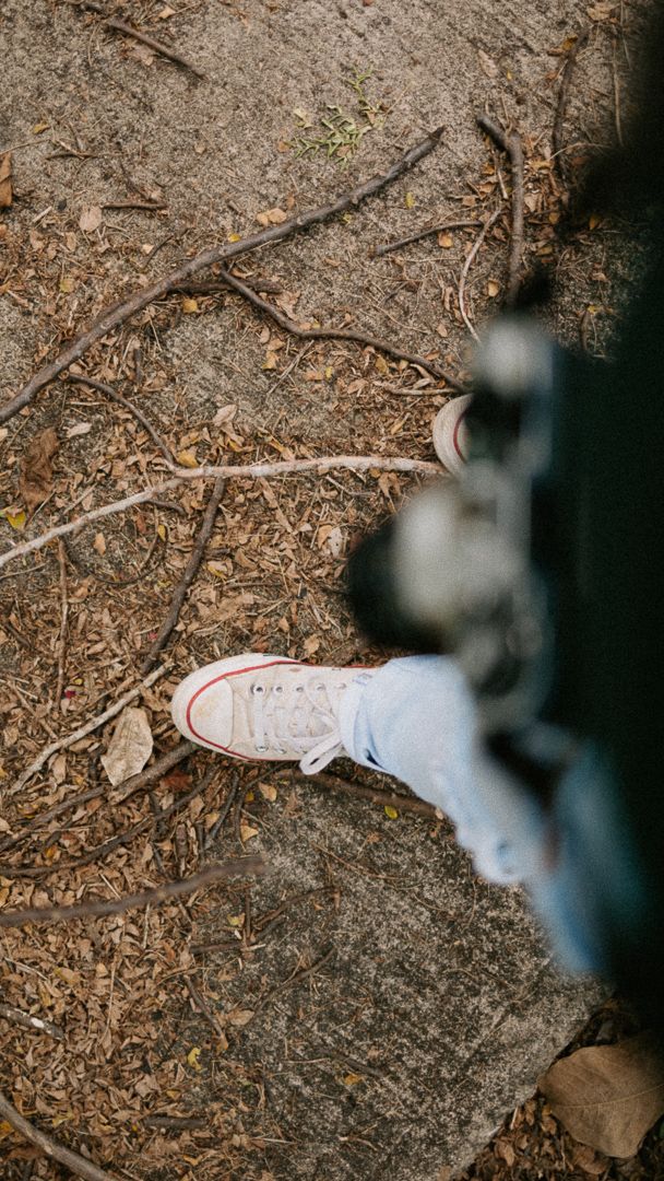 Person wearing white Converse sneakers and jeans standing on rocky ground covered with dry leaves and twigs.
