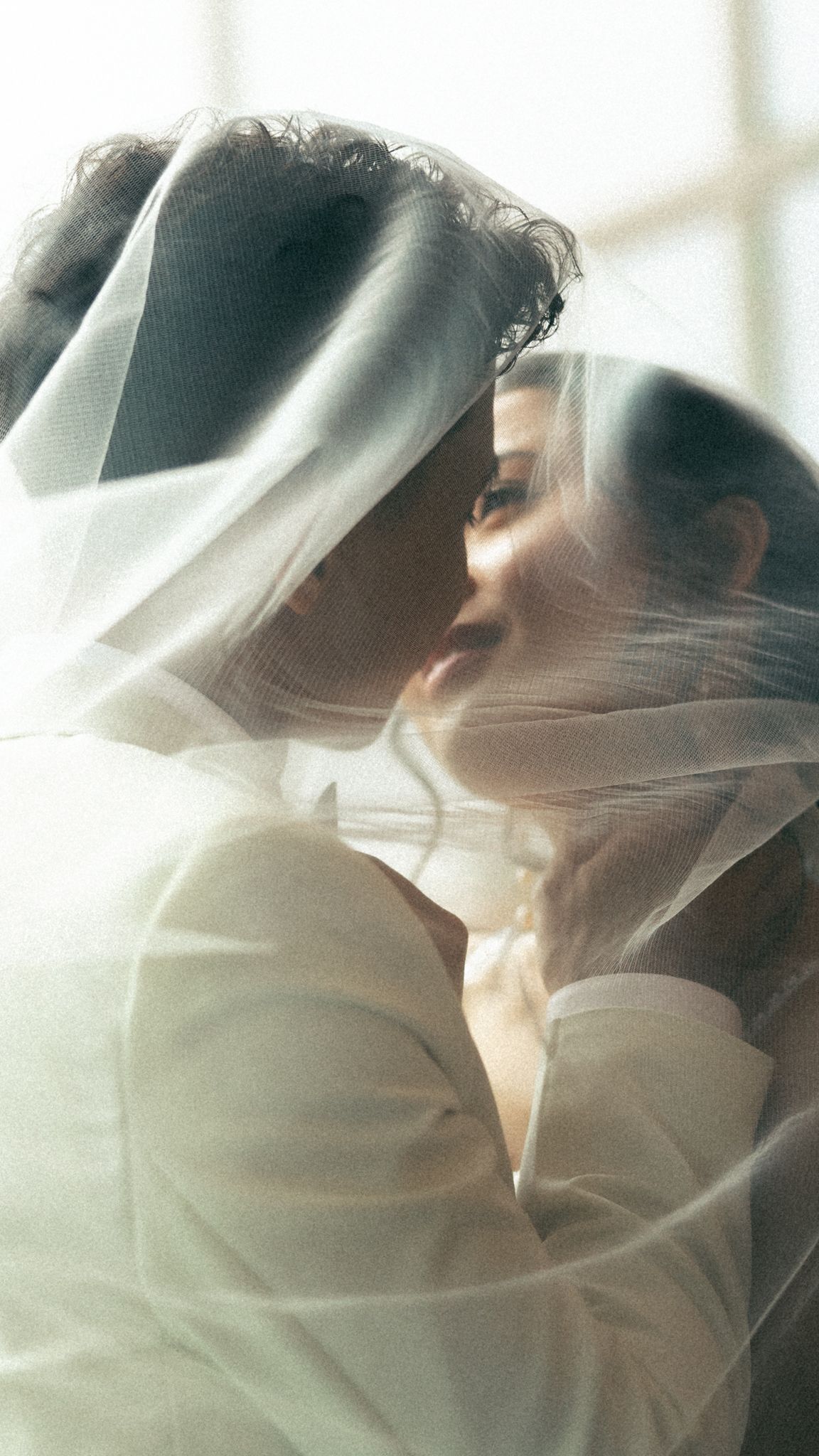 A bride and groom share a romantic kiss beneath a flowing white wedding veil in soft, dreamy light.