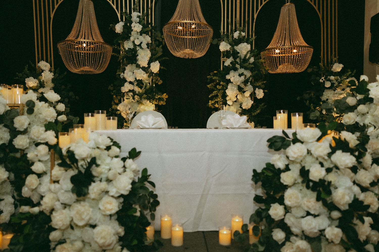 Elegant wedding sweetheart table adorned with white roses, candles, and rattan chandeliers in a romantic candlelit setting.