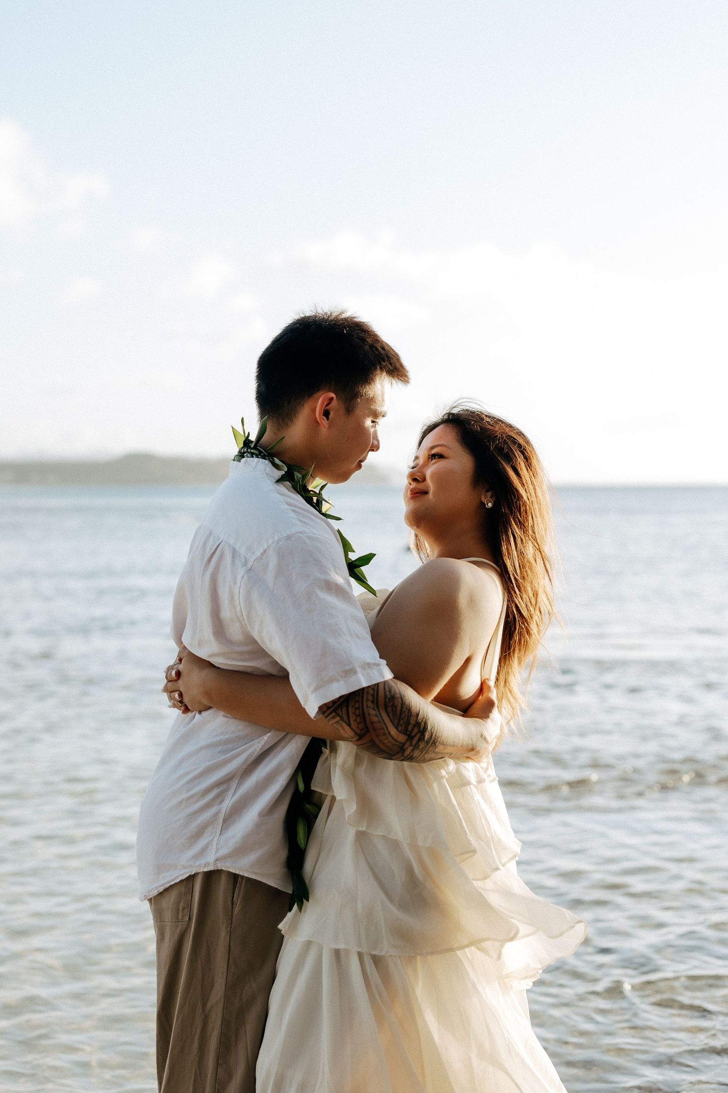 Couple embracing on a beach at sunset, woman in white dress and man in white shirt with floral lei, ocean backdrop.