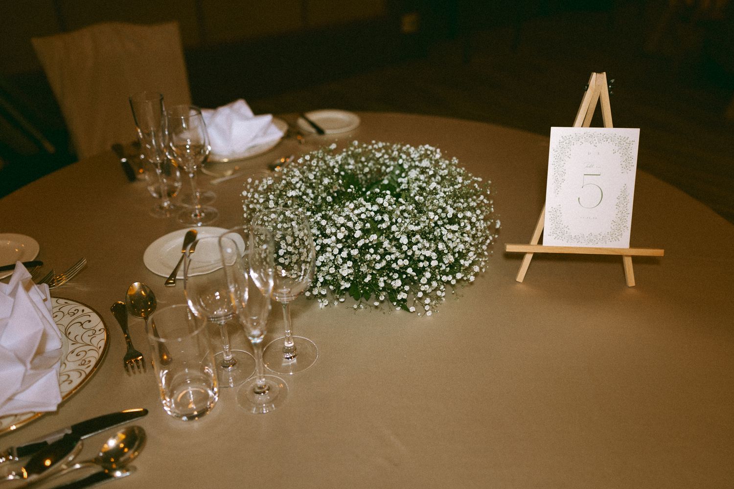 Wedding reception table with baby's breath centerpiece, white napkins, gold cutlery, crystal glasses, and a table number 5 on an easel.