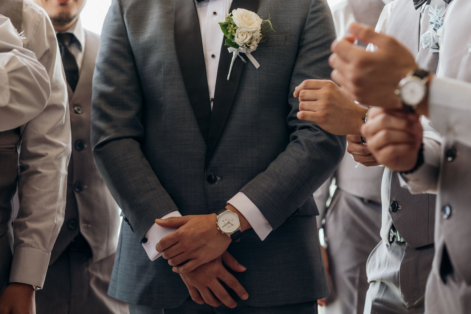 Groom in gray suit with white boutonniere stands with groomsmen in matching attire at a wedding ceremony.