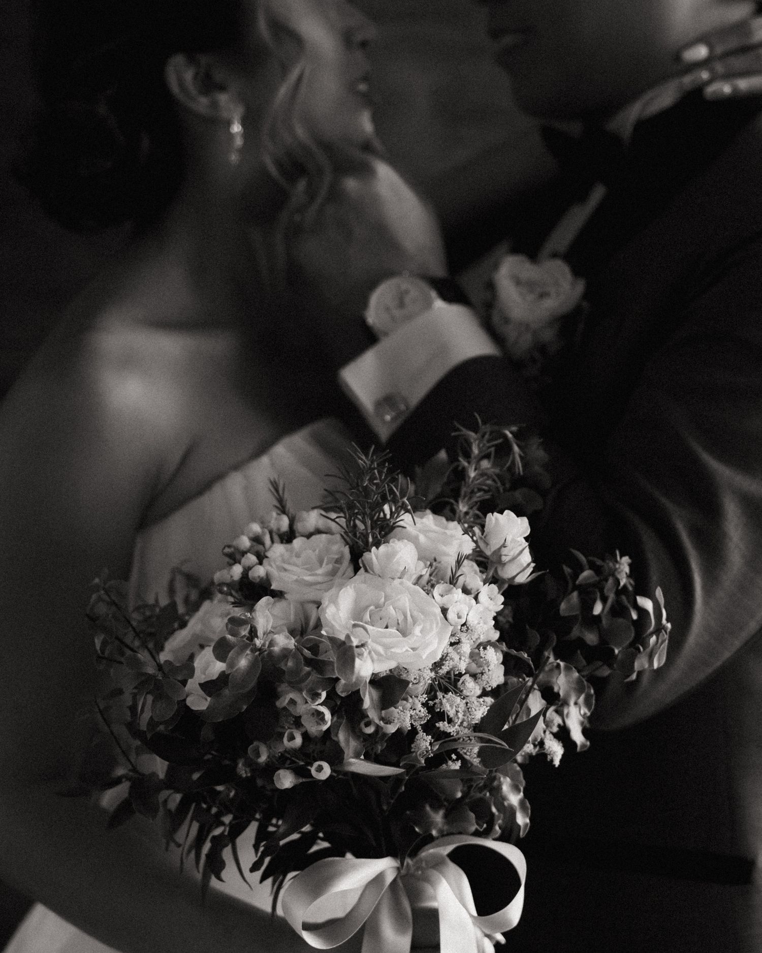 Black and white wedding photo of couple kissing behind a bridal bouquet of roses and flowers tied with ribbon.