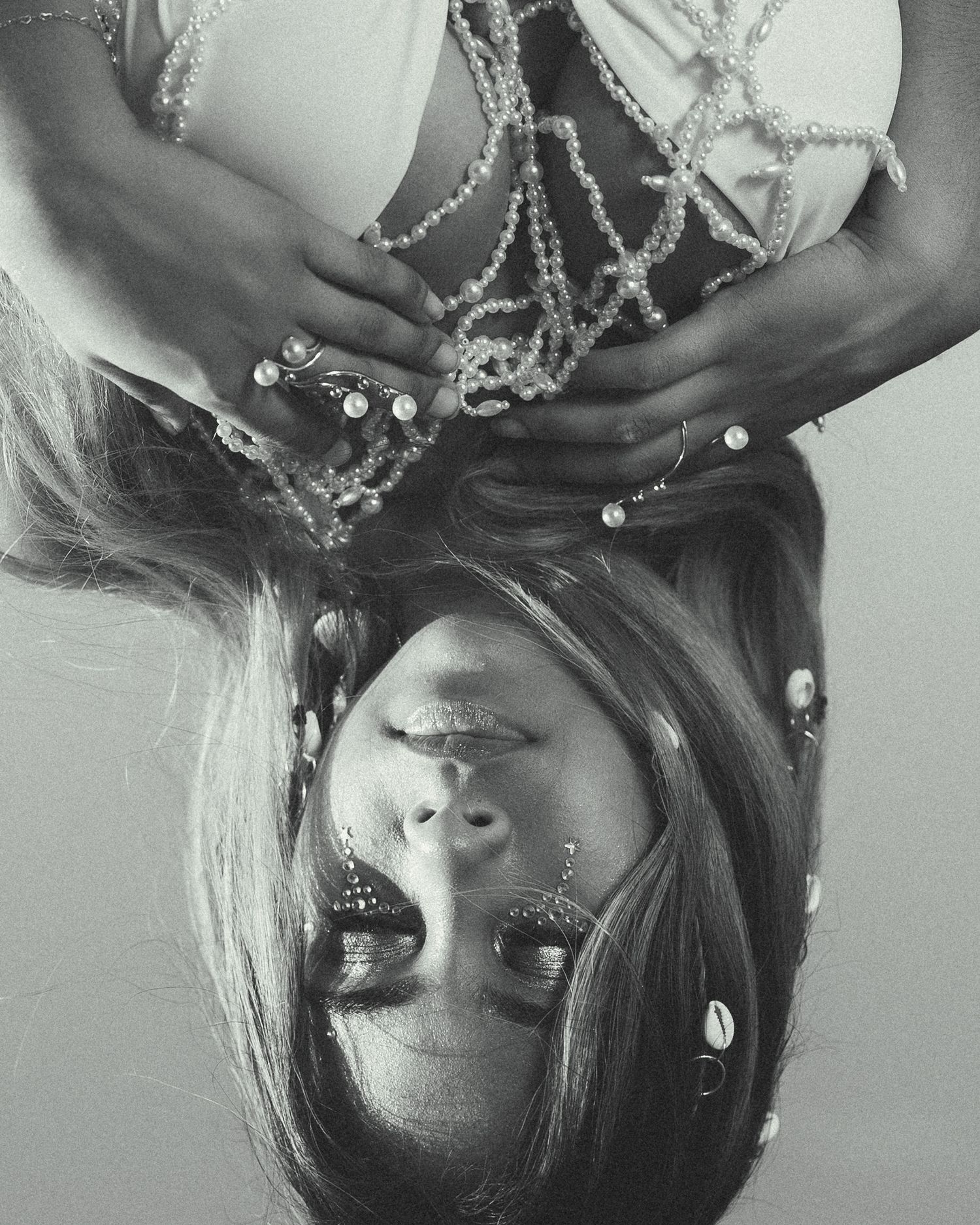 Black and white upside-down portrait of a woman with piercings, adorned with pearl chains and rings.