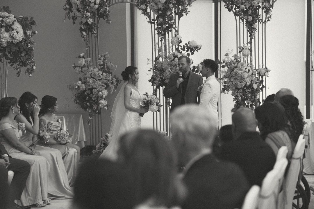 Black and white wedding ceremony photo with bride and groom at floral altar, guests seated in elegant venue.