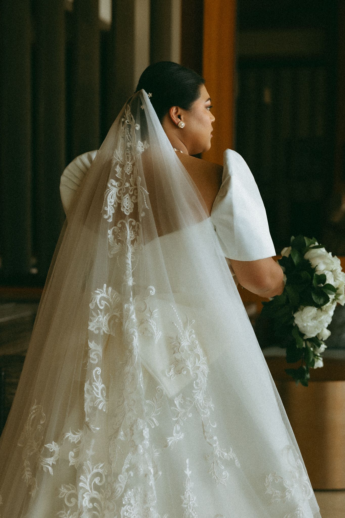 Bride in white lace gown with embroidered veil holds bouquet, viewed from behind in elegant venue.
