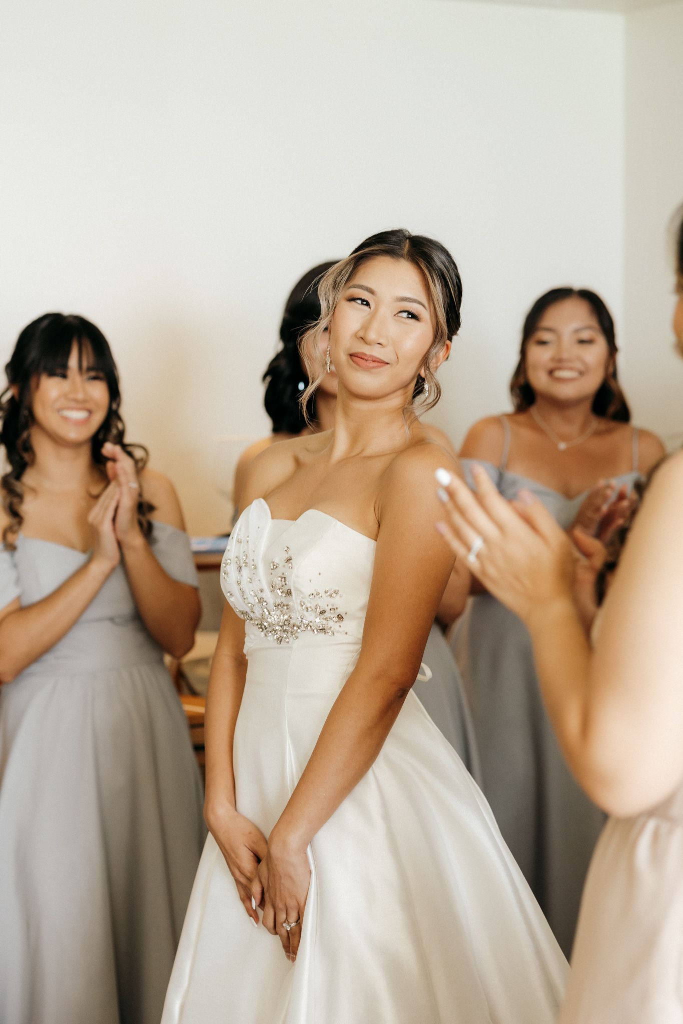 A bride in a strapless white ballgown smiles as her bridesmaids in gray dresses clap and celebrate around her.