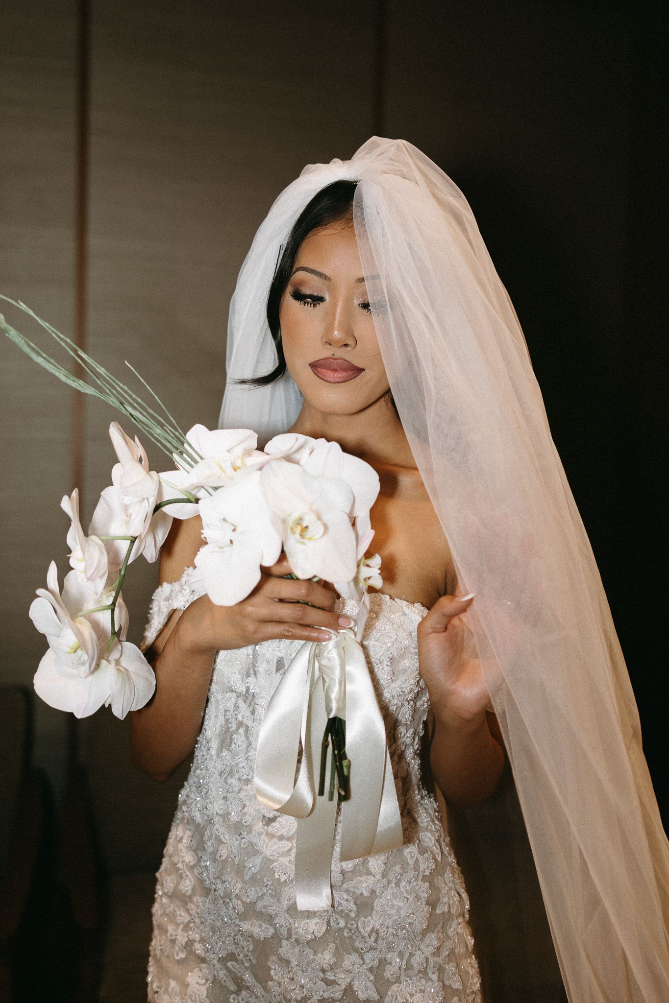 Asian bride in lace gown holding white orchid bouquet with veil draped over shoulder.