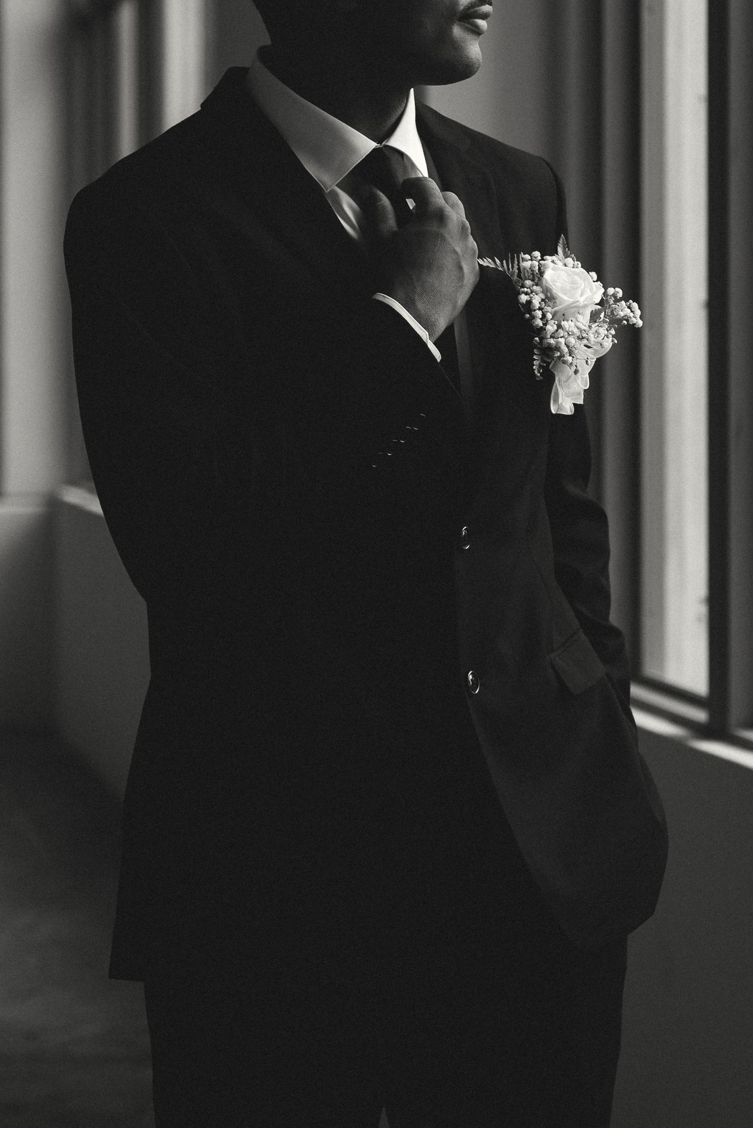 Black and white photo of a groom in a dark suit adjusting his tie with a boutonniere on his lapel.