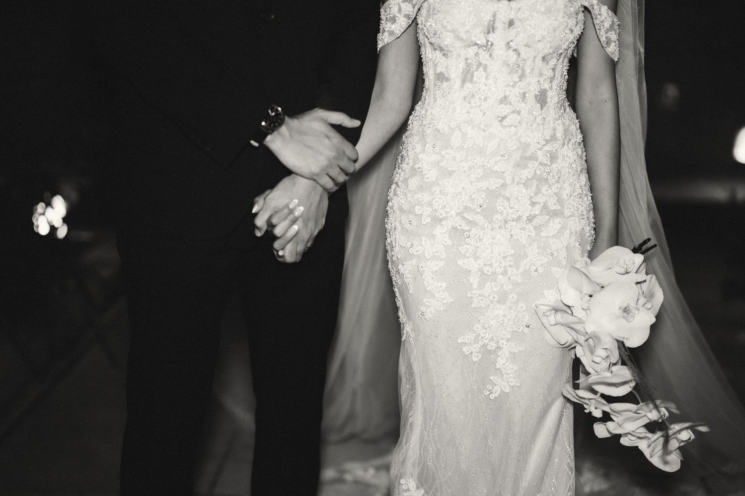 Black and white photo of bride and groom walking together, bride holding floral bouquet.