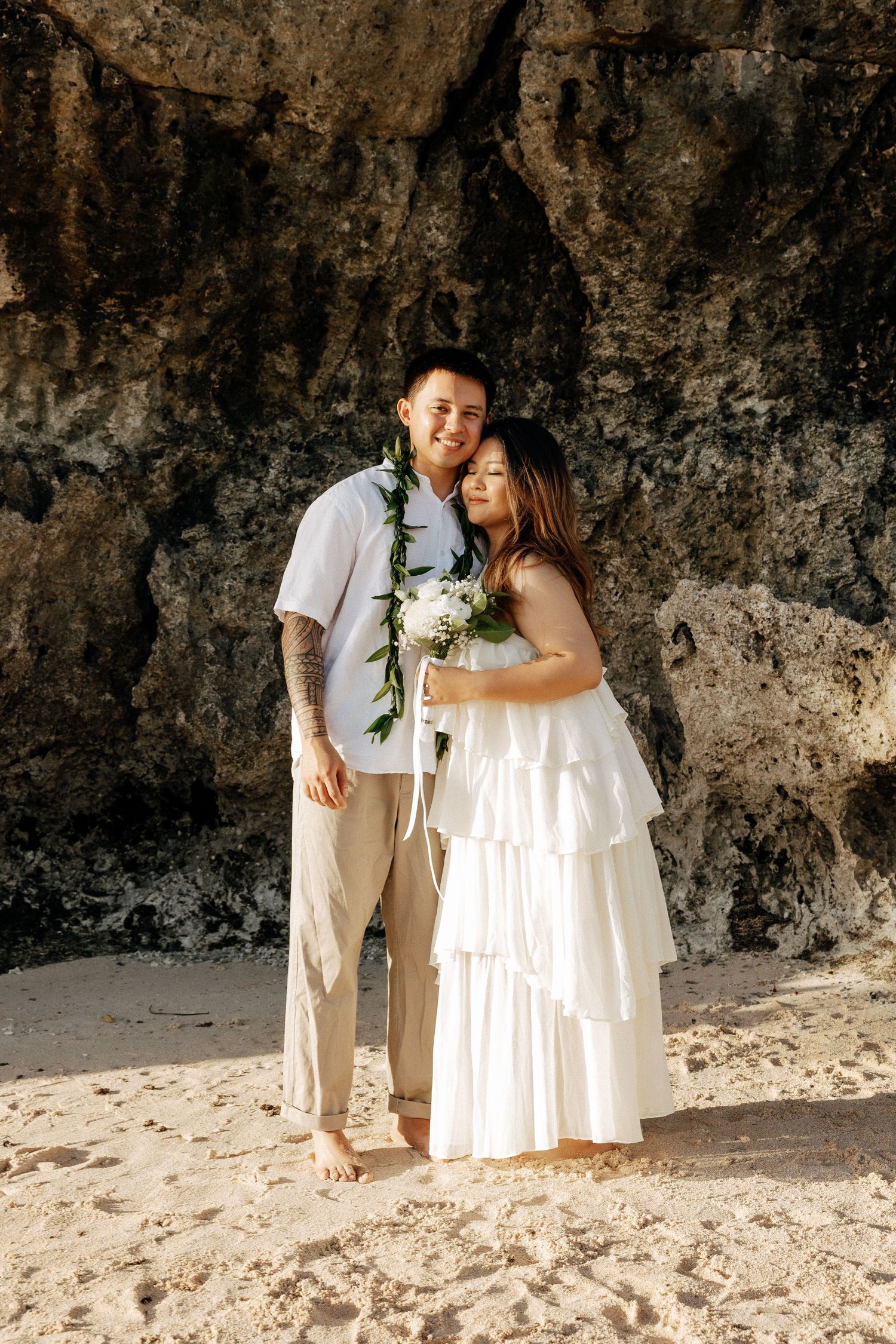 Couple in wedding attire on sandy beach, bride in white tiered dress holding bouquet, groom wearing floral lei.