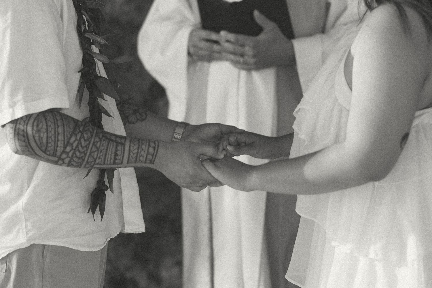 Black and white wedding ceremony photo of groom with tribal tattoos exchanging rings with bride in white dress.