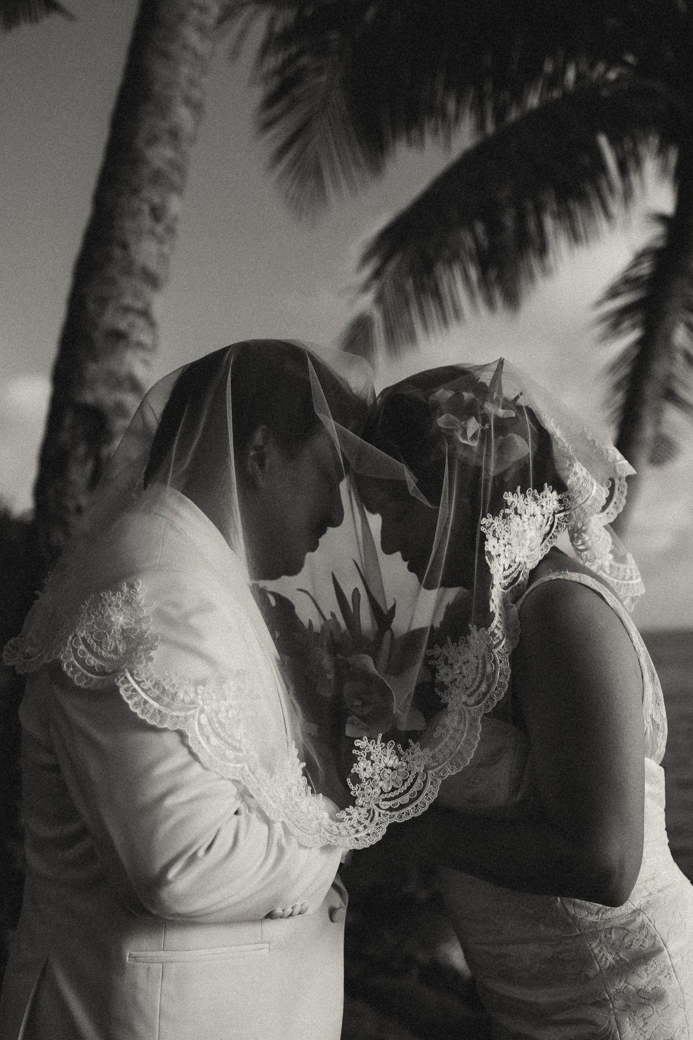 Black and white photo of a bride and groom sharing an intimate moment under a lace veil near tropical palm trees.