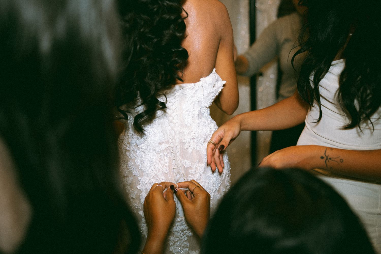 Bridesmaids helping bride in white lace wedding dress, showing henna tattoos on hands in elegant bridal preparation.