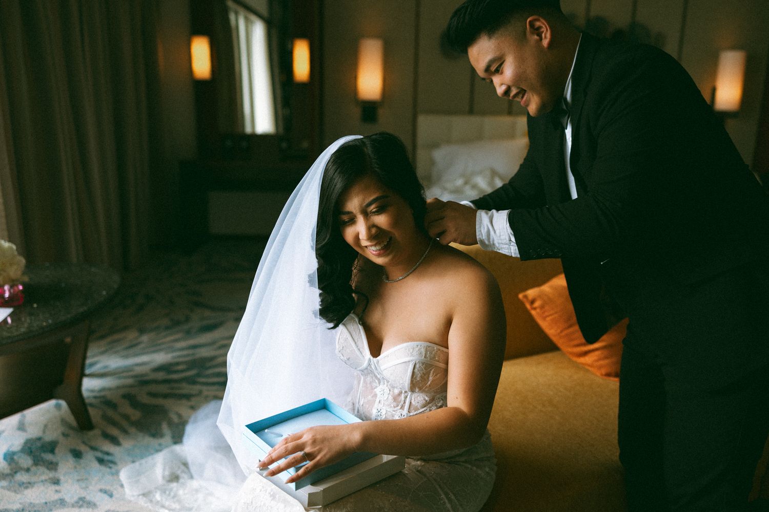 Bride in white strapless gown reads a letter while groom in black tuxedo stands beside her in a hotel room.