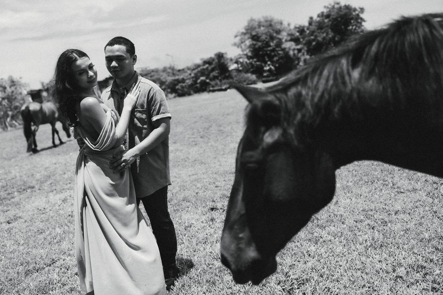 Black and white photo of a couple embracing in a field with a dark horse grazing nearby.