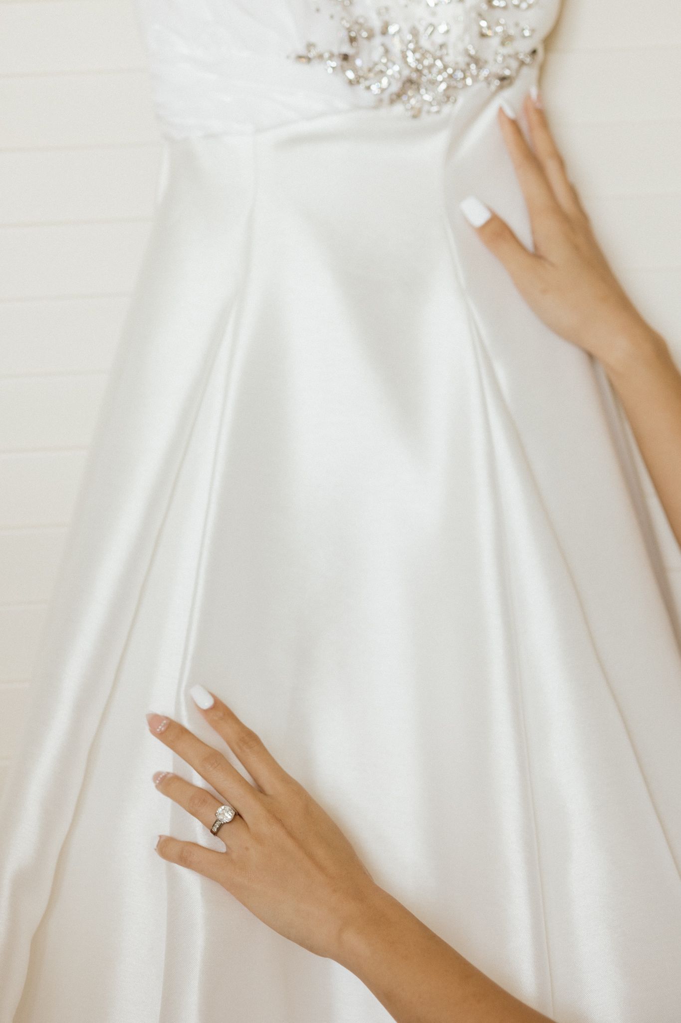 Bride's hands with diamond engagement ring touching elegant white satin wedding dress with crystal beading detail.