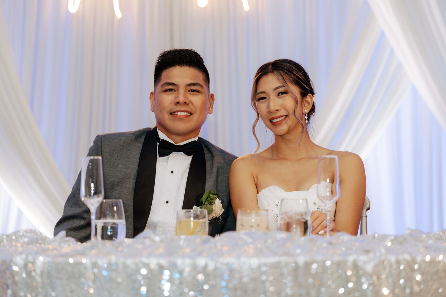 Bride and groom smiling at wedding reception sweetheart table with elegant white drapery backdrop.
