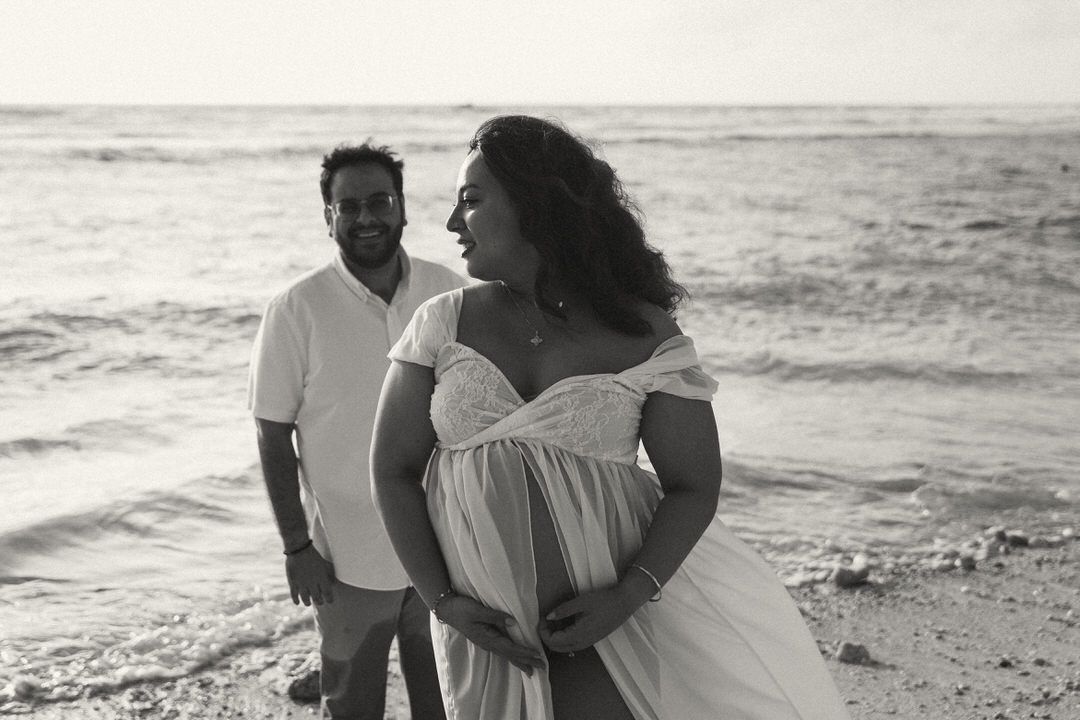 Black and white beach maternity photo of a couple, pregnant woman in flowing white dress, ocean waves behind them.