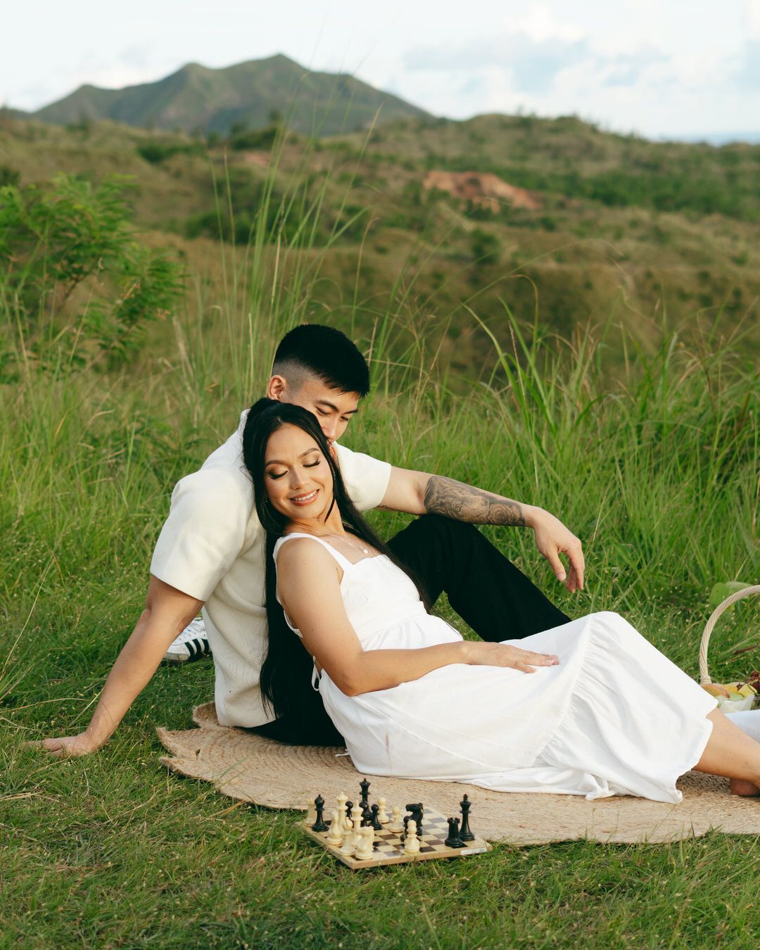 Couple enjoying a romantic outdoor picnic on a blanket with a chess set, surrounded by lush green hills.