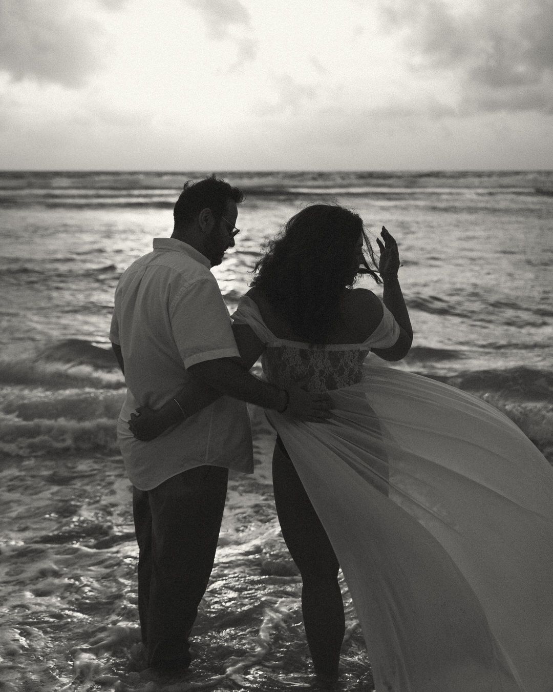Black and white photo of a couple standing in ocean waves at sunset, woman in flowing gown, romantic beach scene.