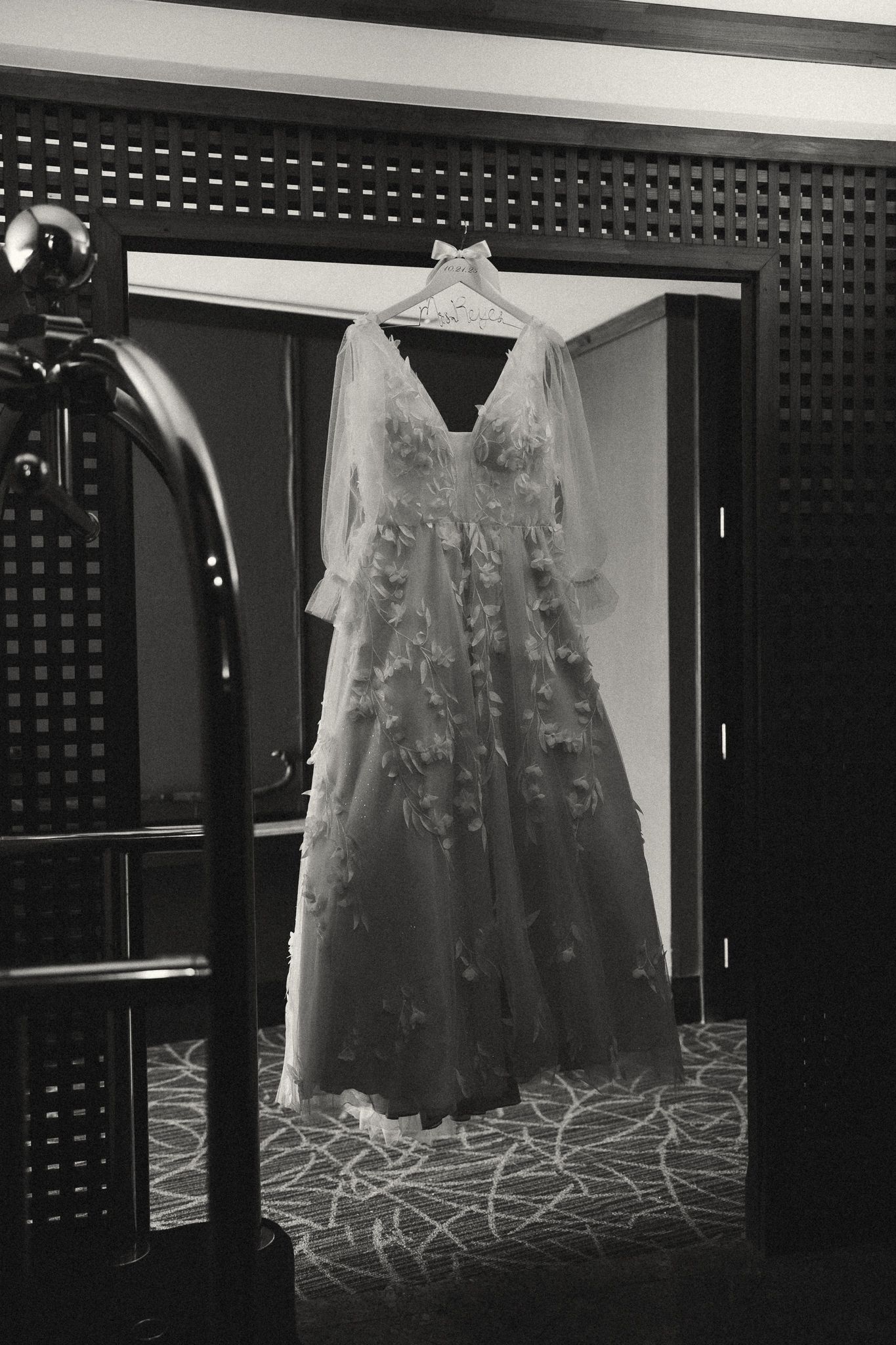 Black and white photo of floral applique wedding gown hanging in doorway of elegant hotel room.