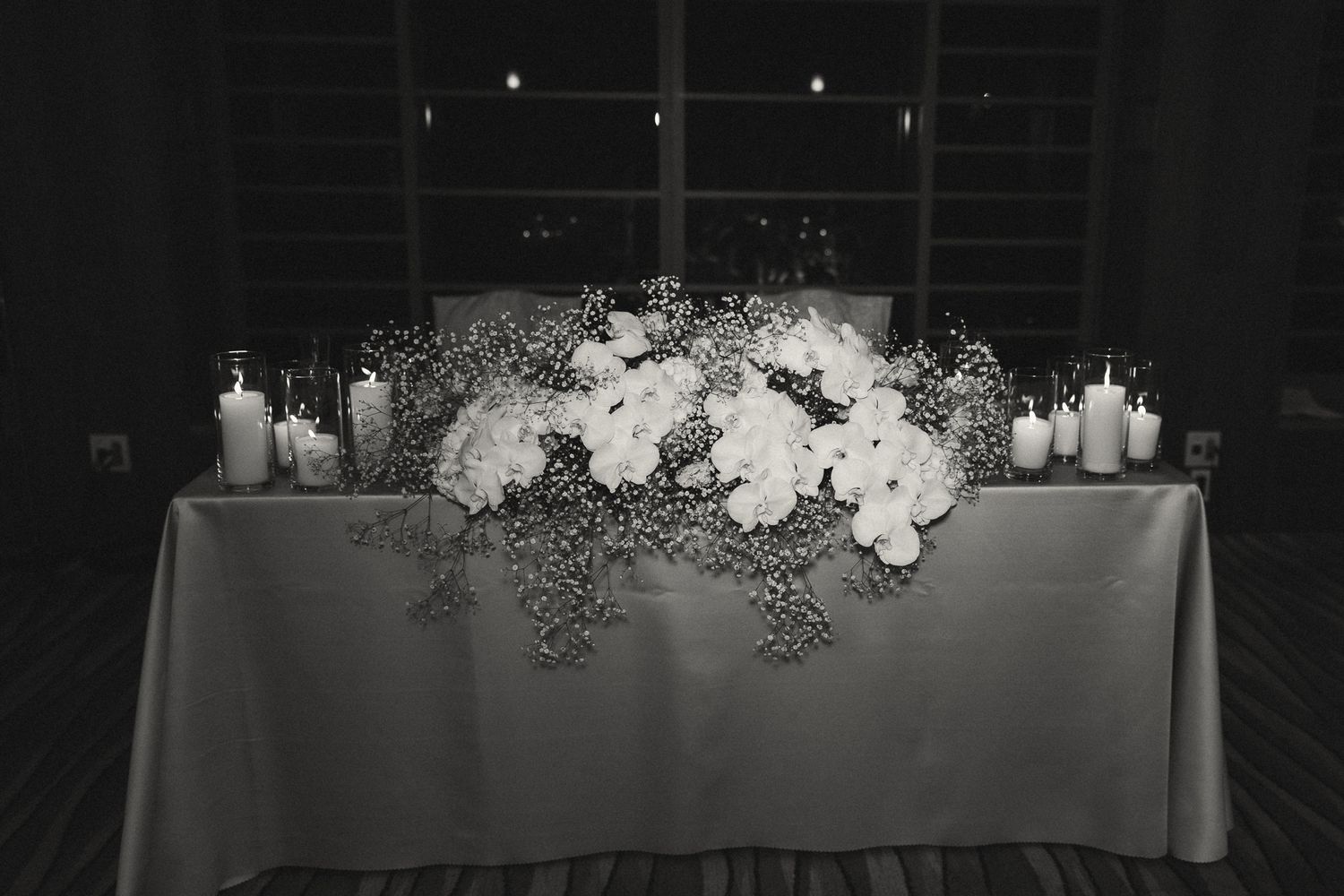Black and white photo of elegant wedding sweetheart table with white floral centerpiece and candles.