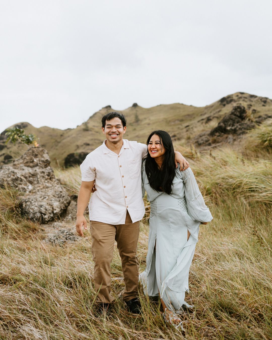 A smiling couple walks through a grassy hillside, the woman in a light blue dress and man in a white shirt and tan pants.