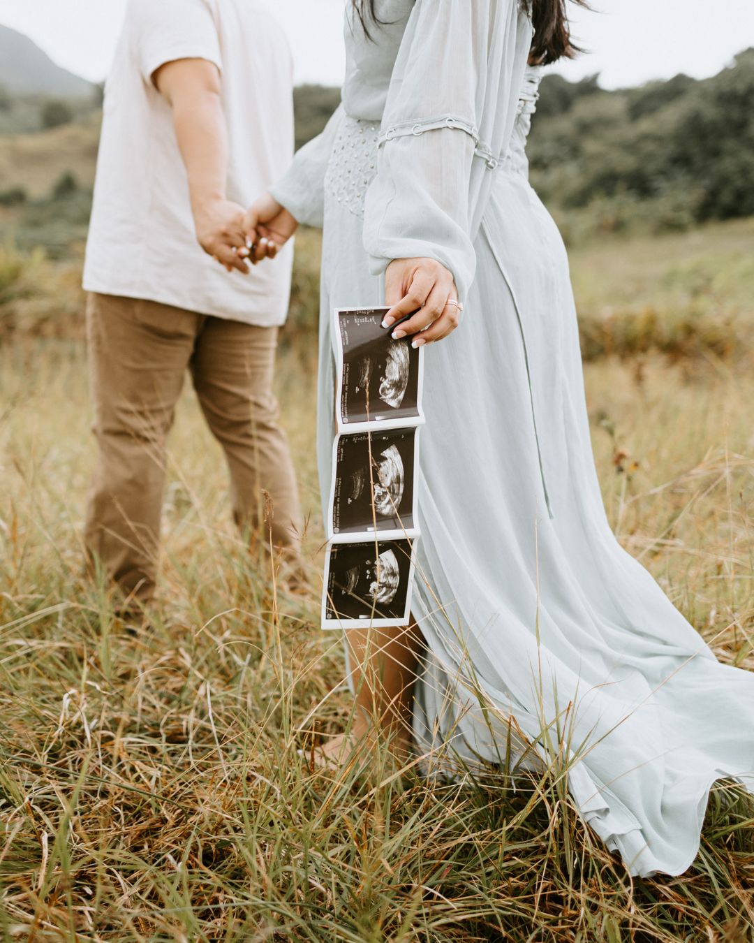 Couple holding hands in a field, woman in light blue dress displaying ultrasound photos, pregnancy announcement outdoors.