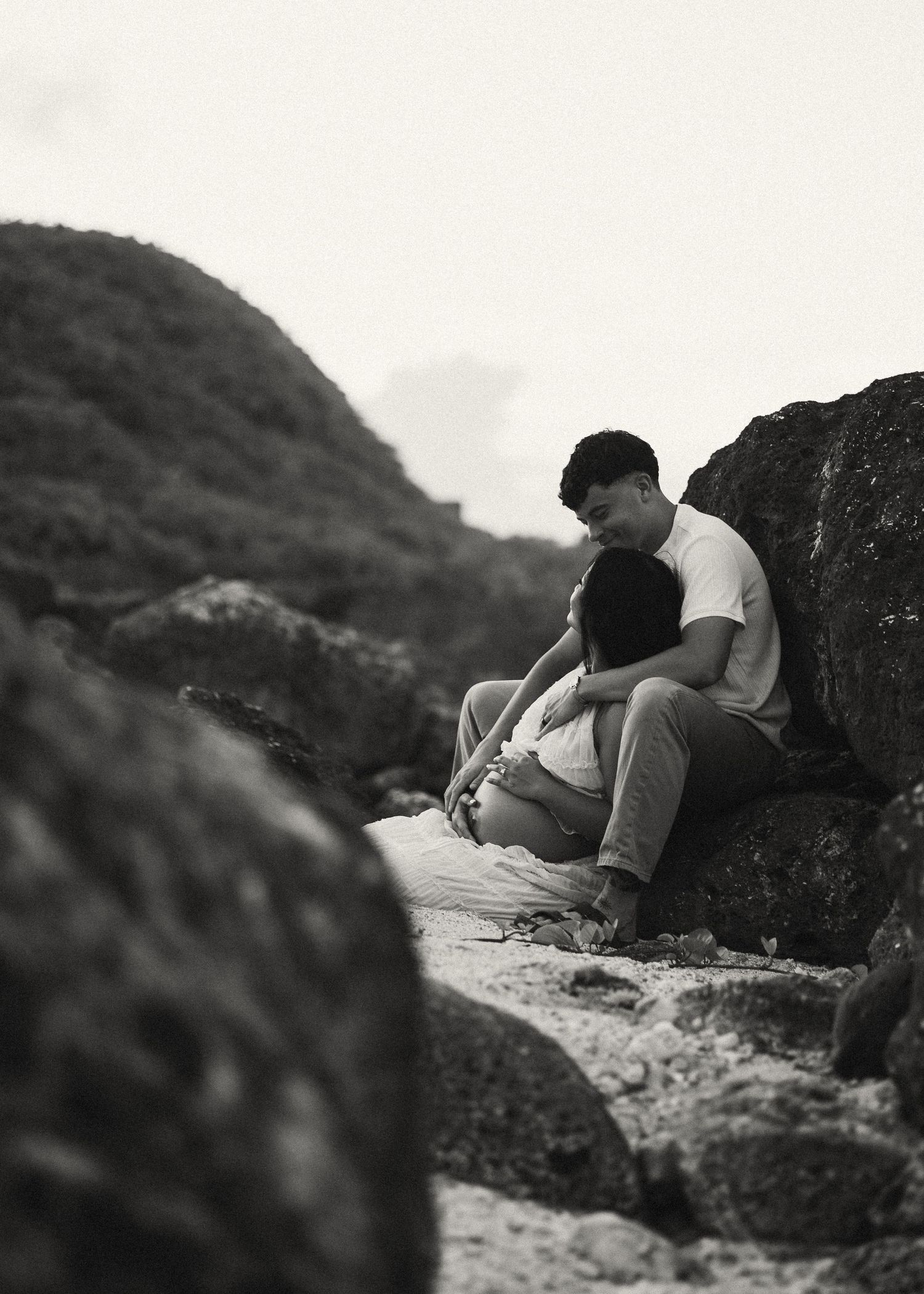 Black and white photo of a couple sitting together on rocky beach, sharing an intimate moment surrounded by large boulders.