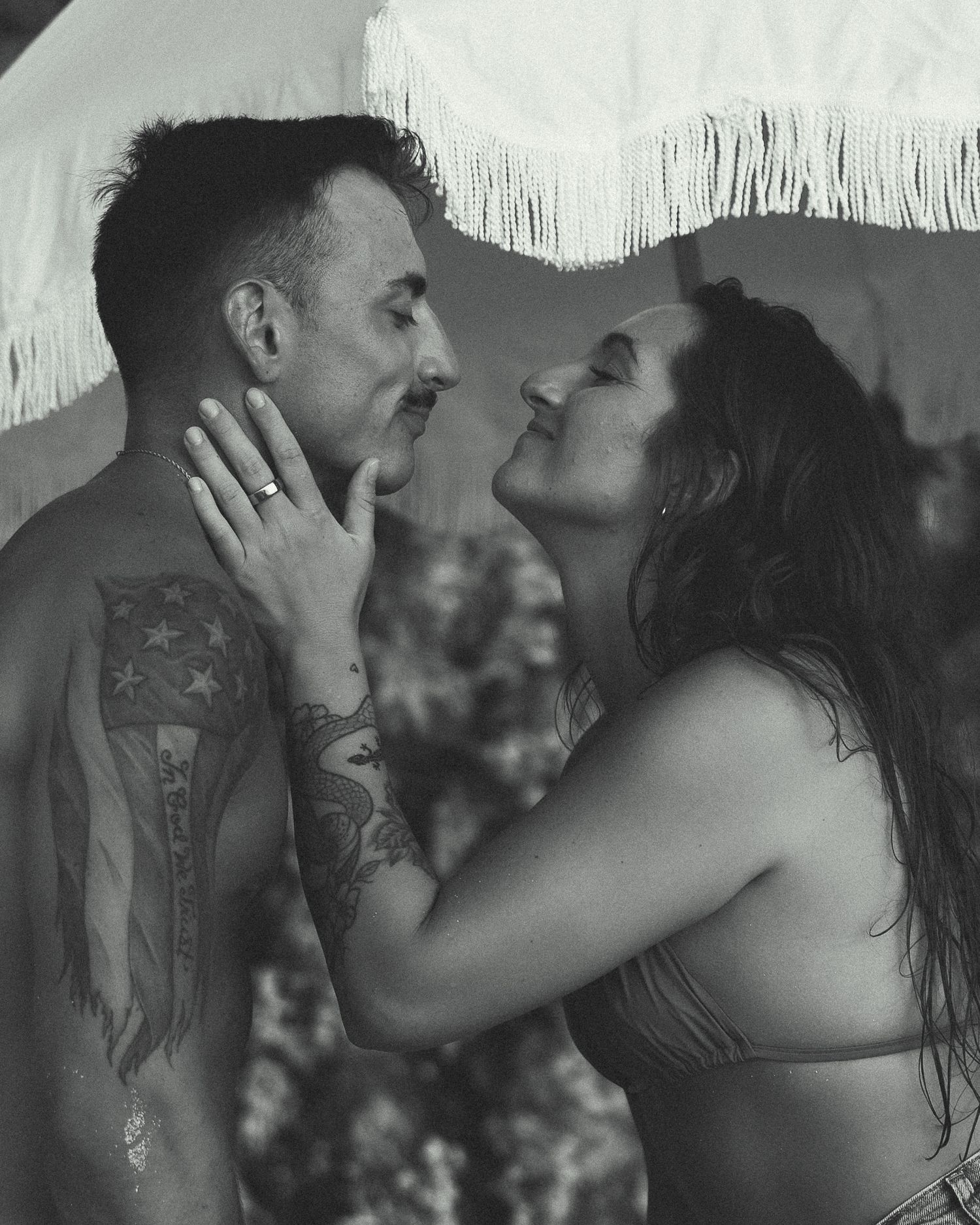 Black and white photo of a tattooed couple sharing an intimate moment outdoors under a fringed umbrella.