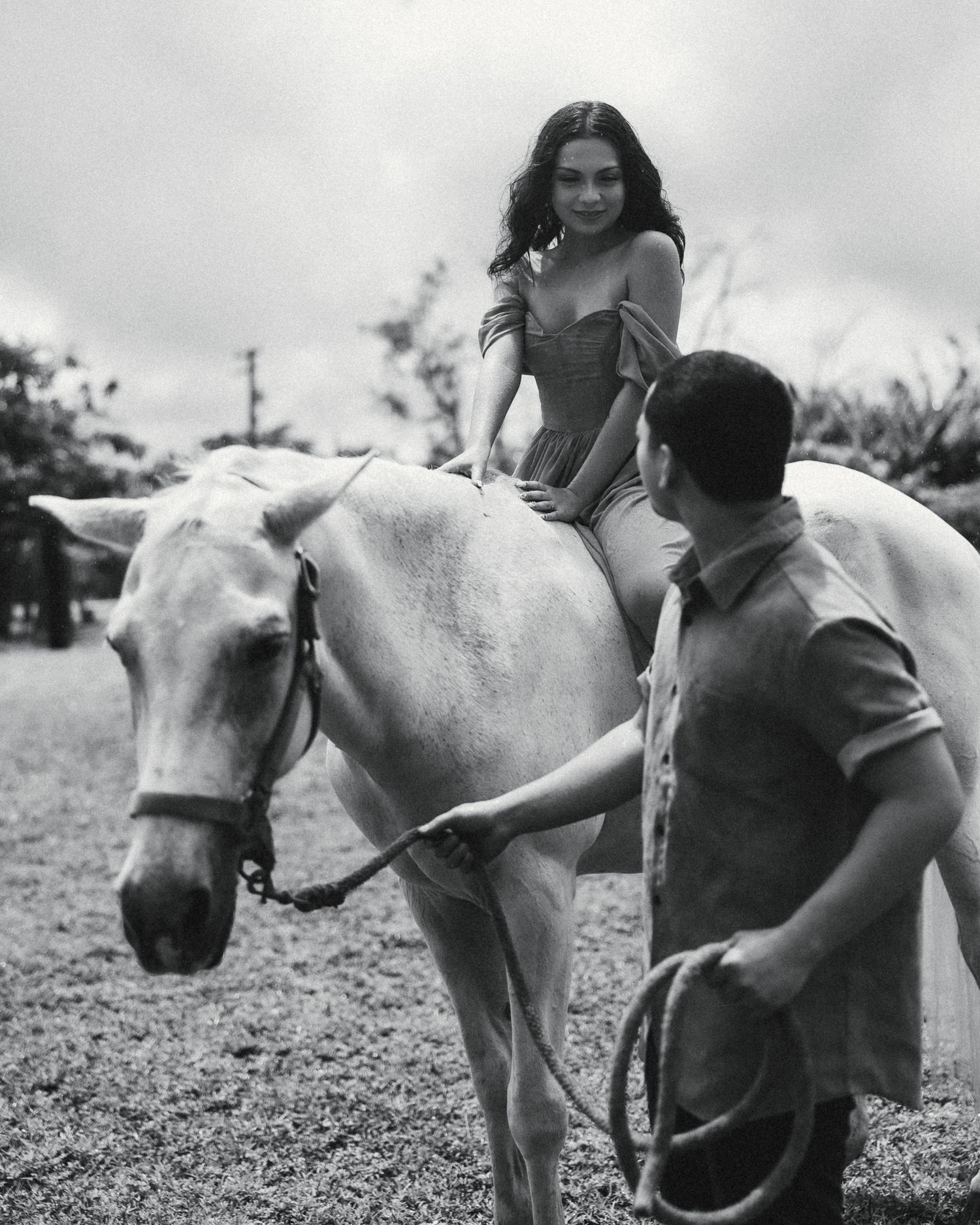 Black and white photo of a woman in a dress sitting on a white horse, held by a man on a grassy field.