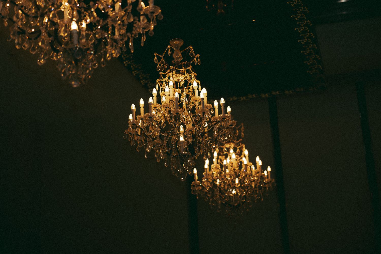 Three ornate crystal chandeliers with glowing candle lights hang from a dark ceiling in an elegant room.