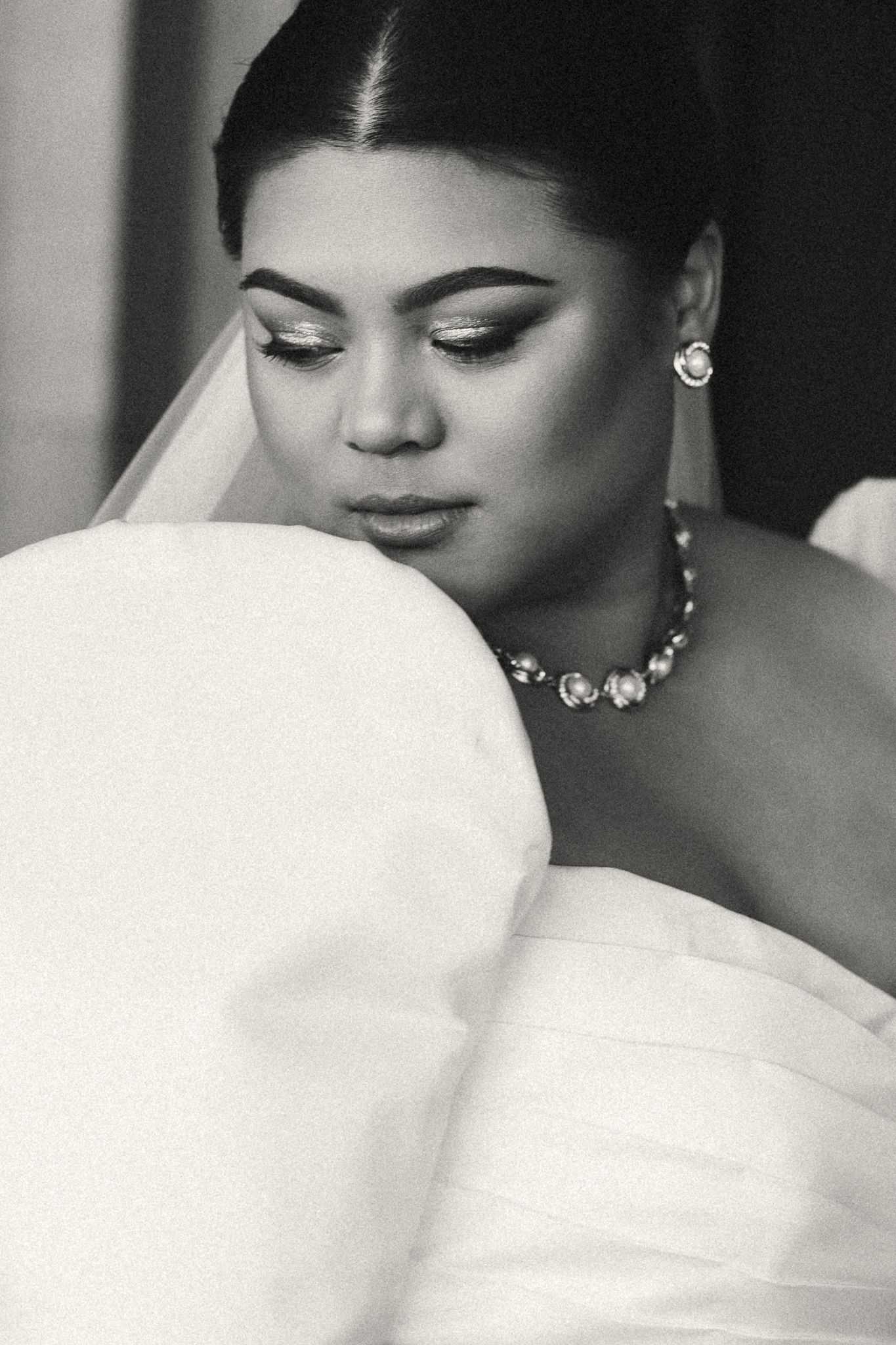 Black and white portrait of a bride in a white gown and veil, wearing a crystal necklace and earrings, looking down.