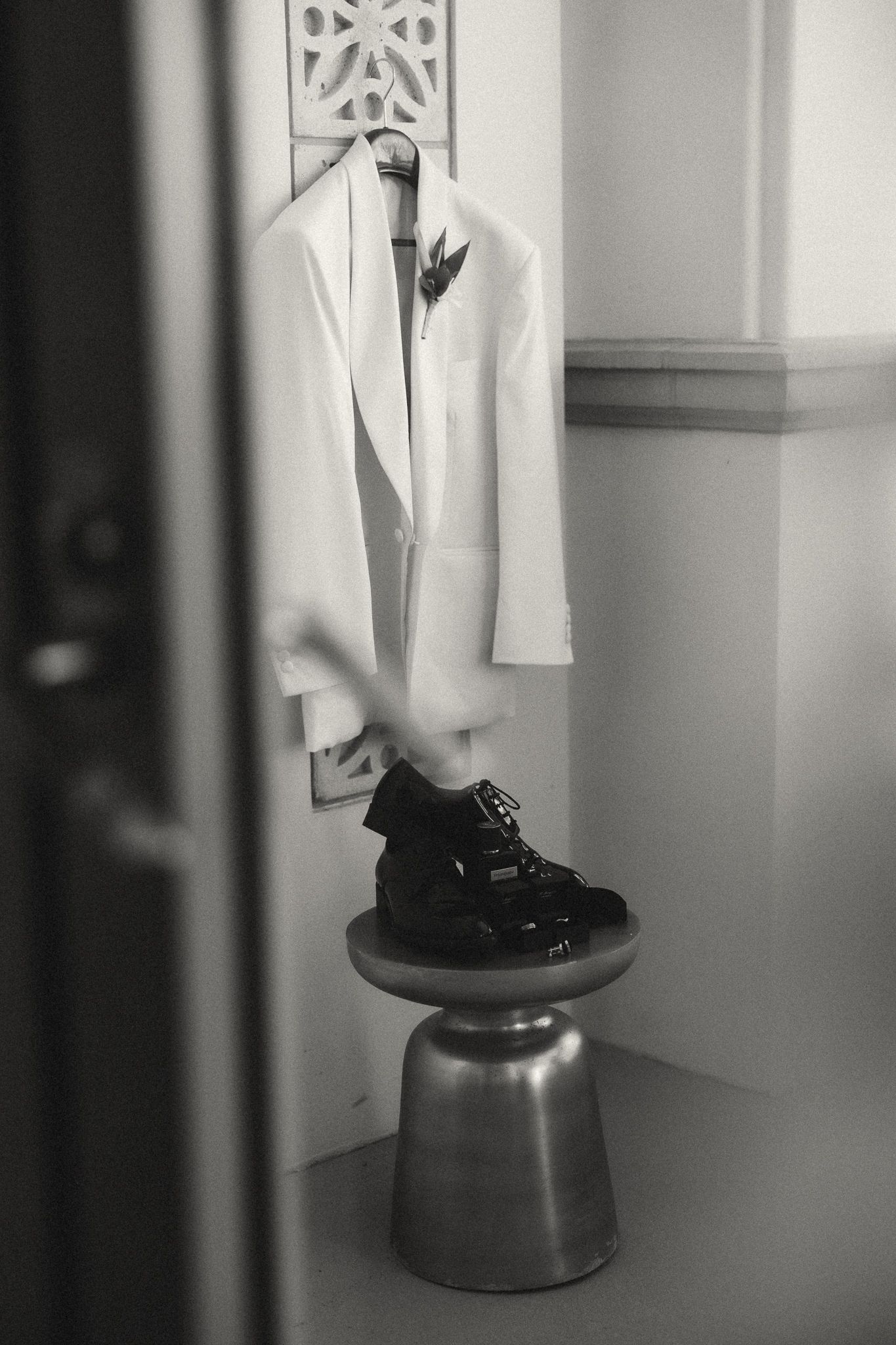 Black and white photo of a white wedding suit jacket hanging on a wall above black dress shoes on a silver stool.