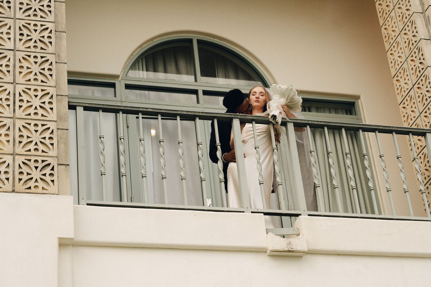 Woman in white outfit standing on a balcony holding a black hat and white bouquet on a sunny day.