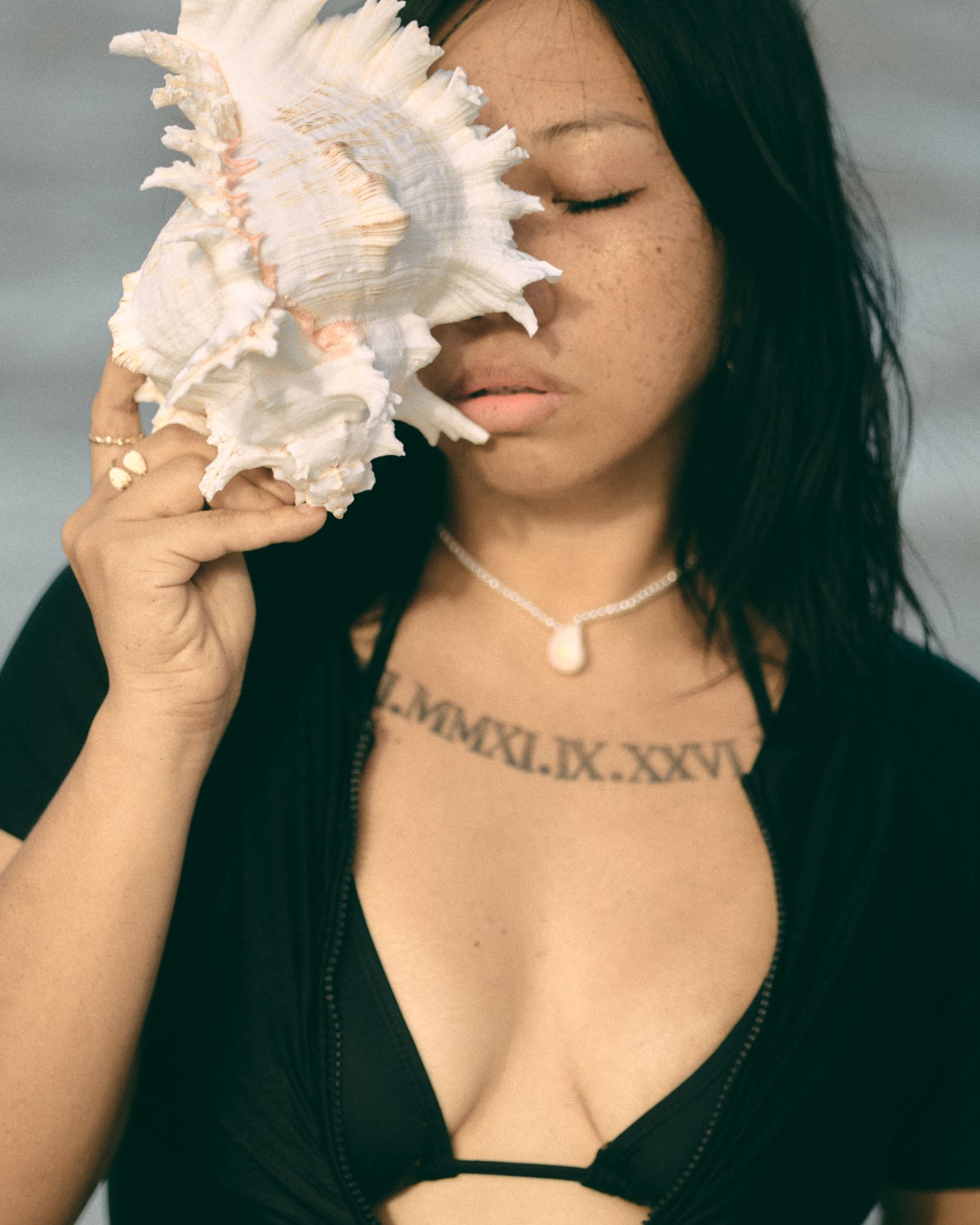 Woman holding large conch shell near face, wearing black bikini top and pearl choker necklace with Roman numeral chest tattoo.
