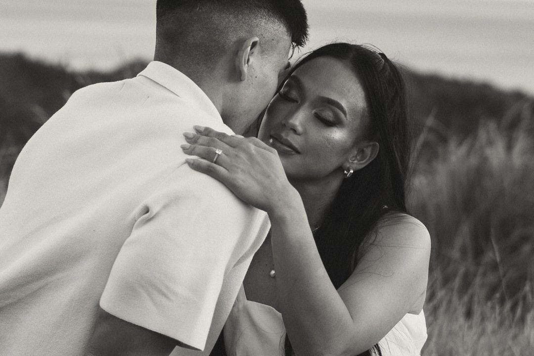 Black and white photo of a couple embracing romantically in a grassy field, the woman smiling softly at her partner.