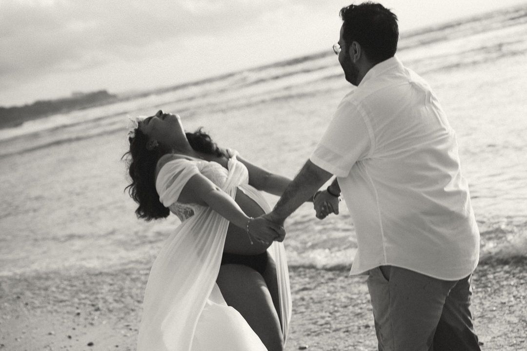 Black and white photo of a couple dancing and holding hands on a beach by the ocean waves.