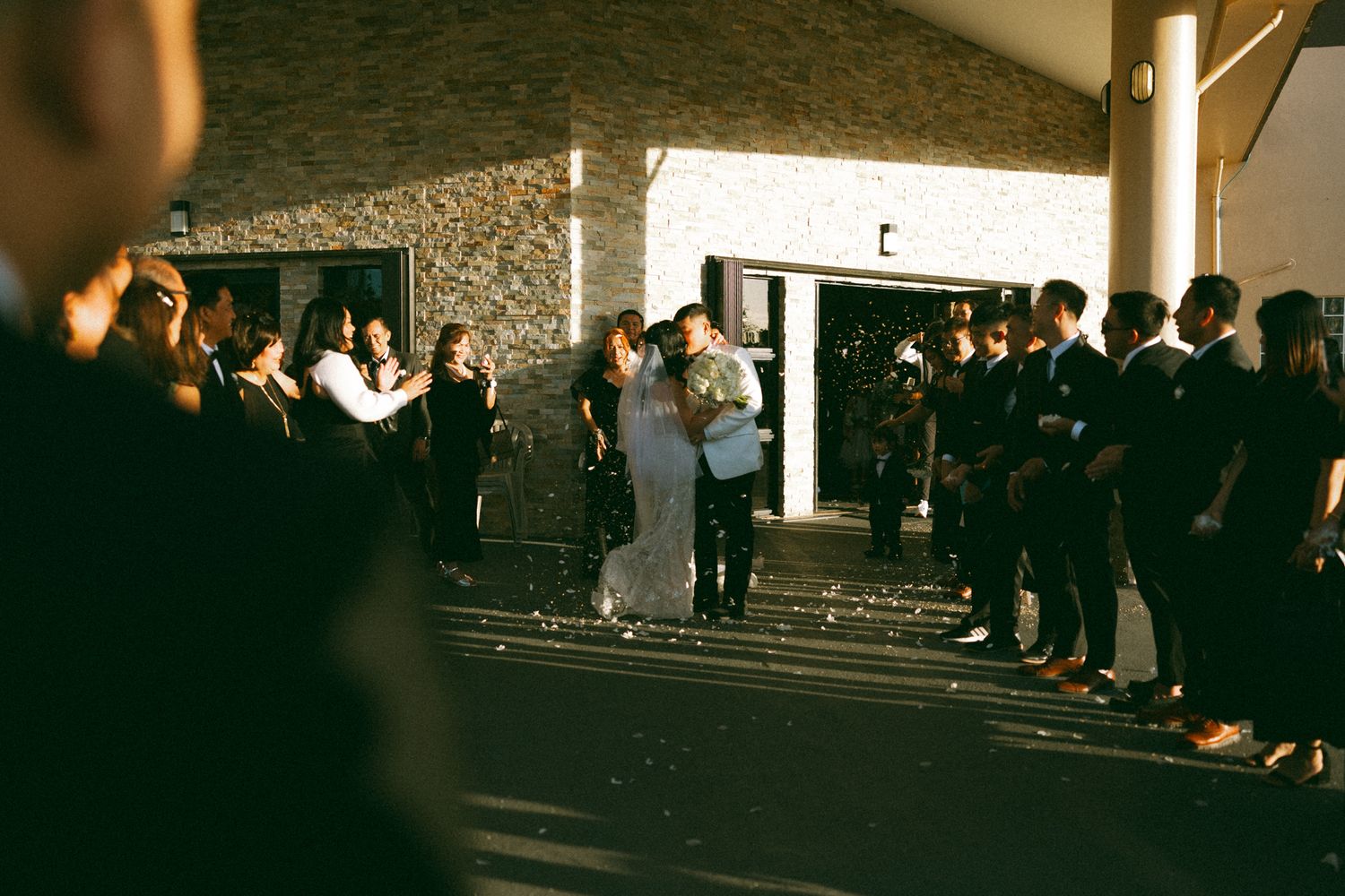 Wedding couple kissing outdoors surrounded by guests celebrating after ceremony at a modern venue.