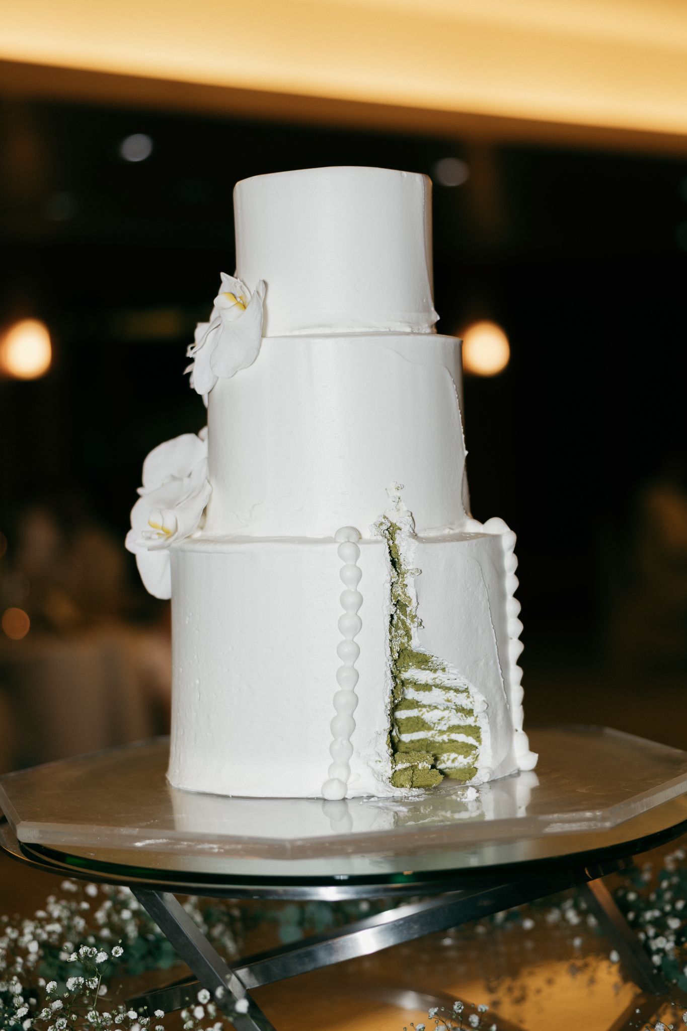 Elegant three-tier white wedding cake with white orchids and pearl details on a glass stand.