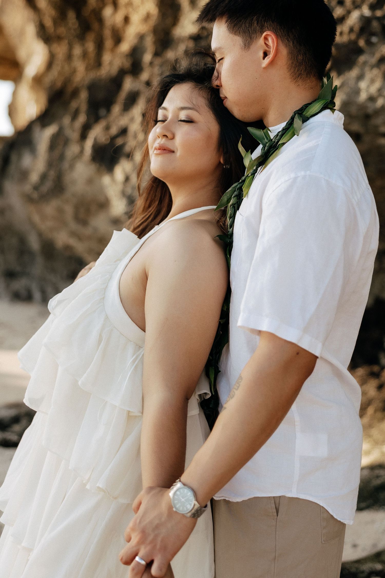 Couple in white attire sharing a romantic moment on a rocky beach, man wearing a green lei kissing woman's head.
