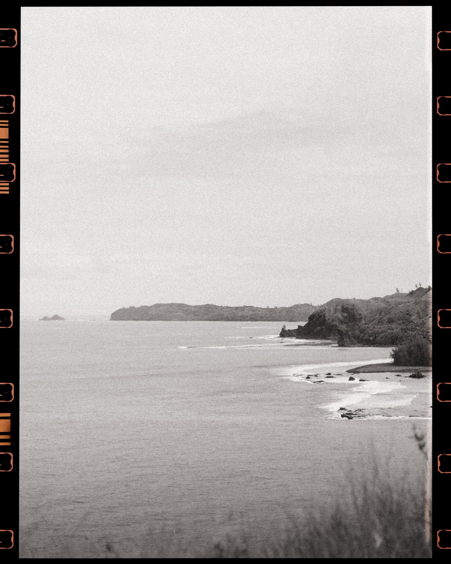 Black and white film photo of a coastal landscape with calm ocean waters, rocky shoreline, and lush green headland under overcast skies.