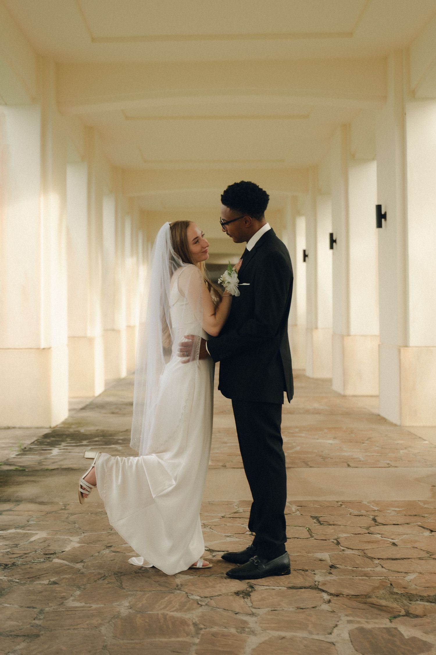 Interracial couple in wedding attire sharing a romantic moment in a sunlit colonnade hallway.