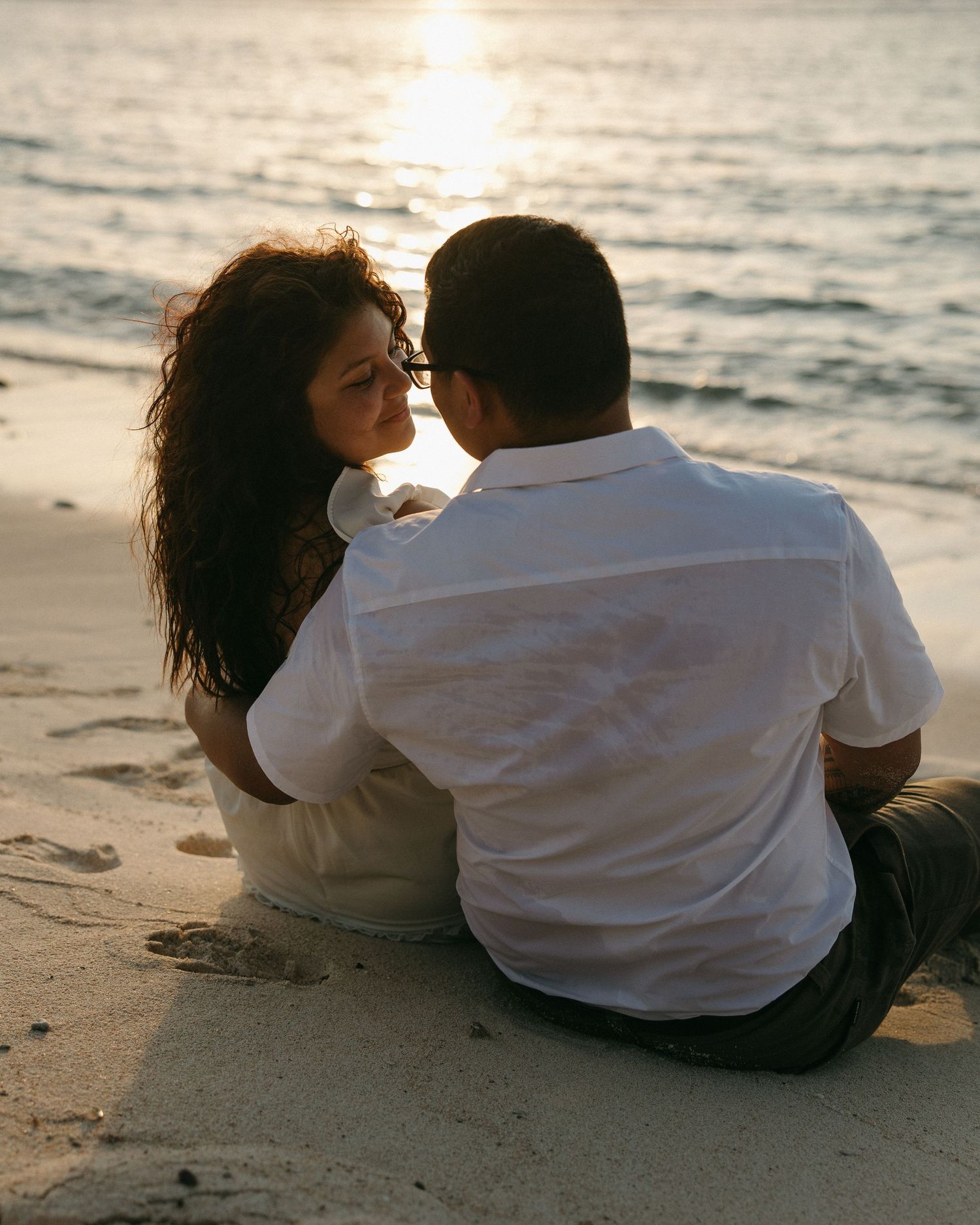 Romantic couple sitting together on a sandy beach at sunset, sharing an intimate moment by the ocean waves.