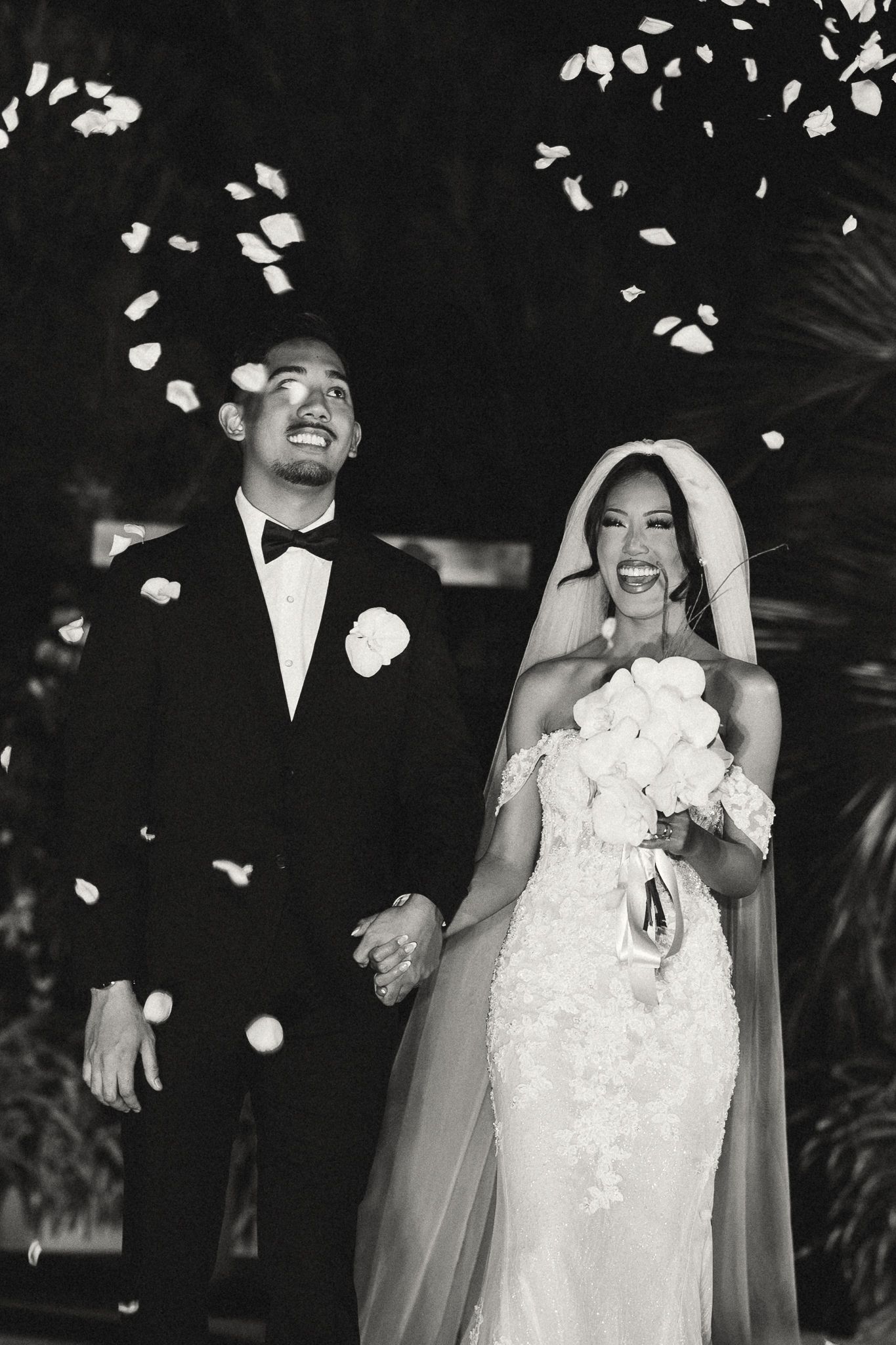 Black and white photo of a joyful bride and groom walking down the aisle as flower petals fall around them.