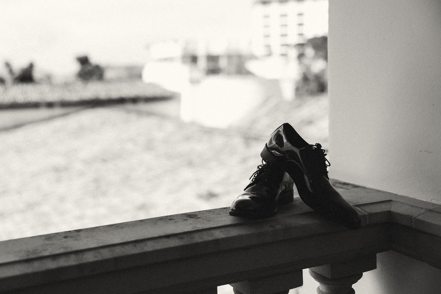 Black and white photo of men's dress shoes resting on a balcony railing with a blurred beach scene in the background.