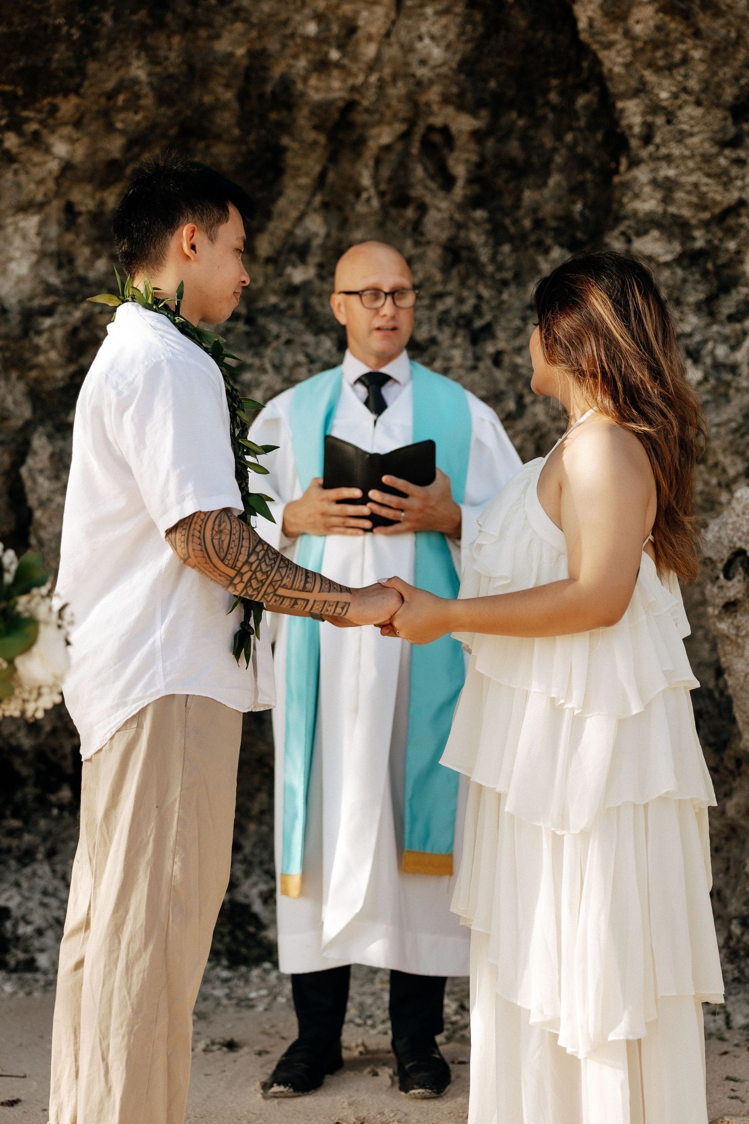 Couple exchanging vows at a beach wedding ceremony with an officiant holding a book, rocky cliffs in the background.