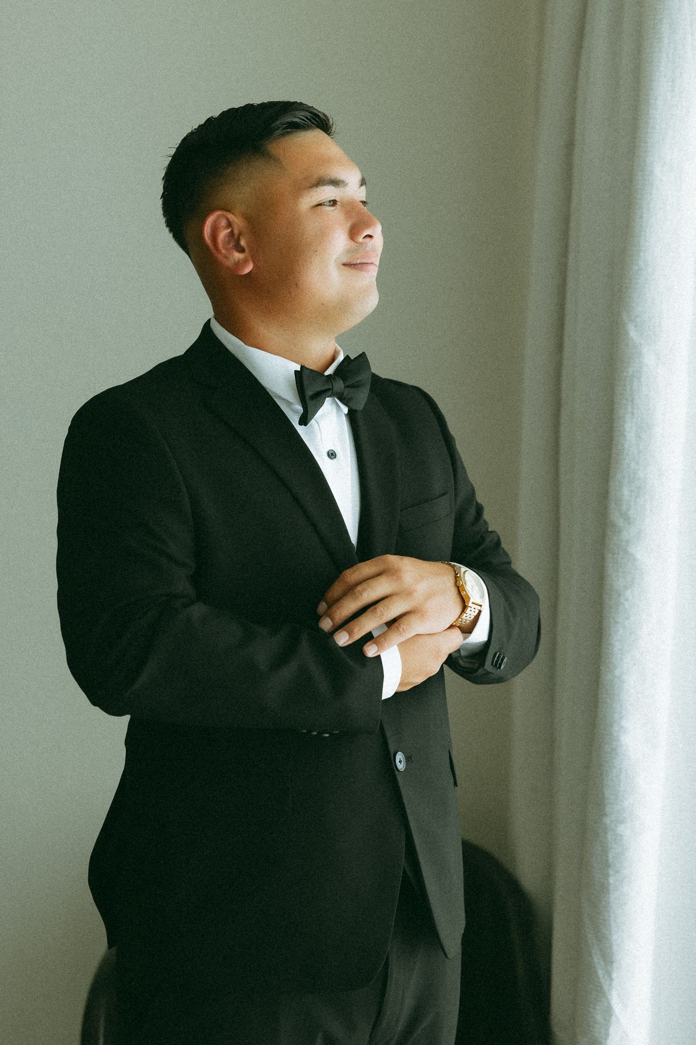 A groom in a black tuxedo with bow tie adjusts his cufflinks while gazing upward near a window.