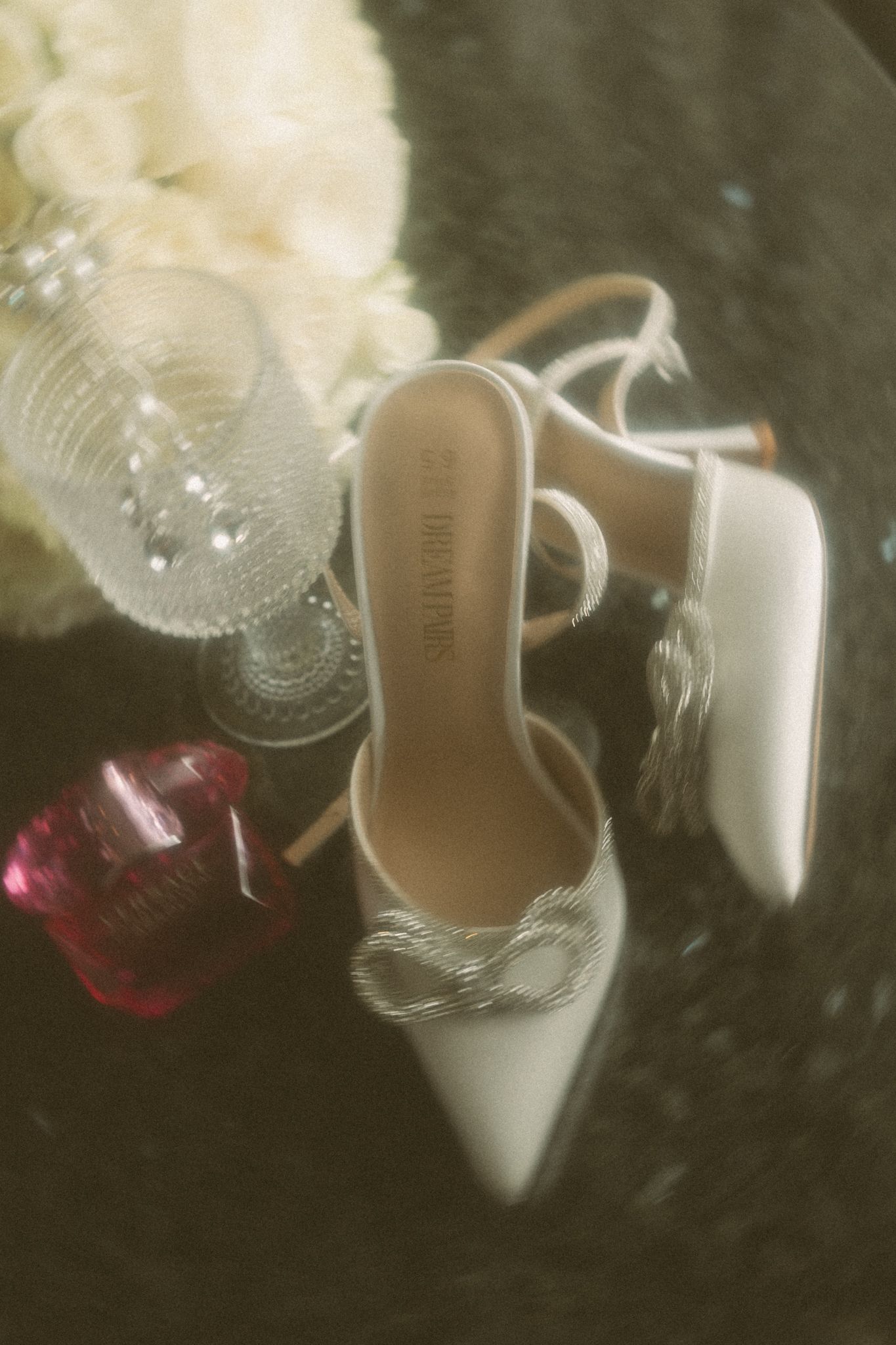 White bridal heels with silver bow detail beside crystal glassware and pink perfume bottle on dark surface.